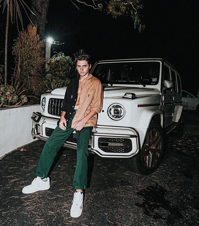 Isaak Presley in a black-orange shirt and green pants sitting in front of his luxurious vehicle.